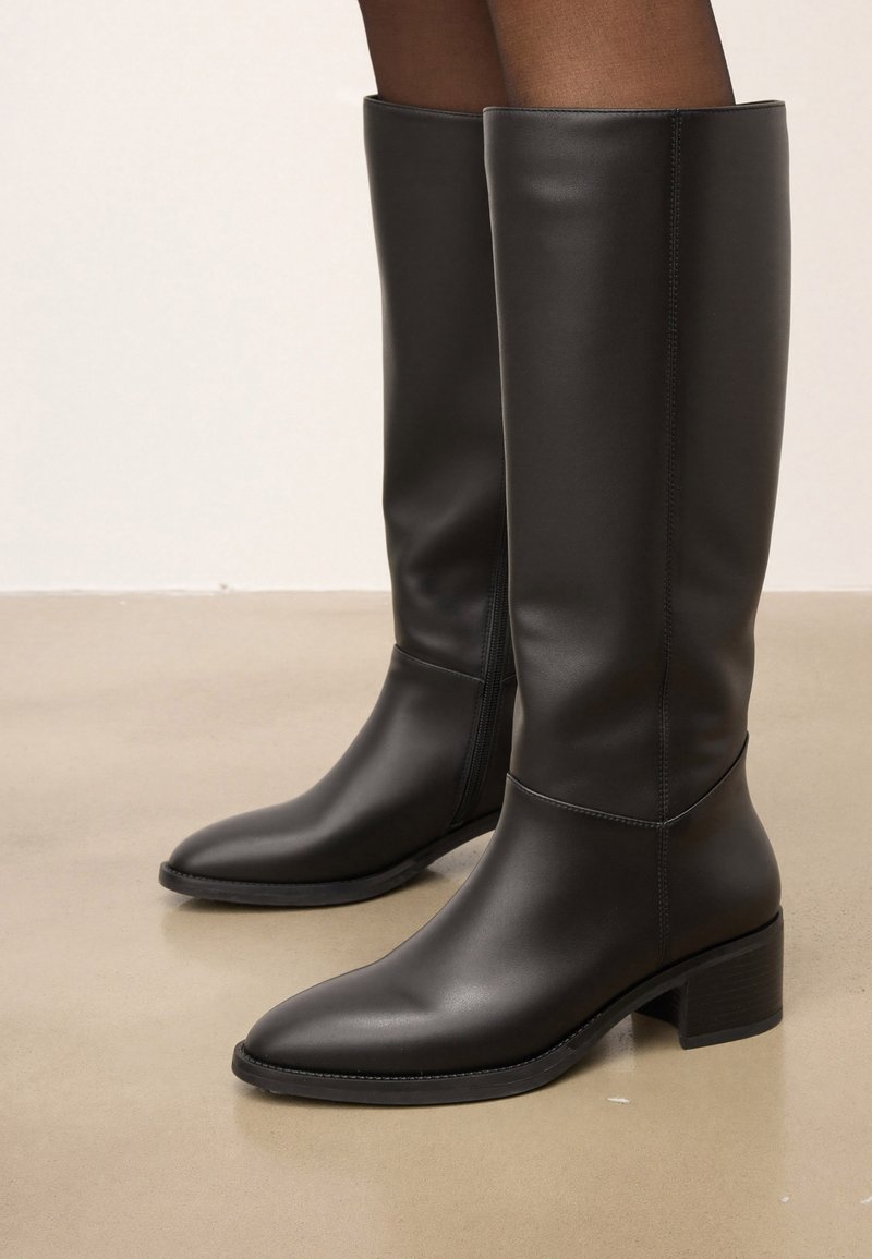 Botas de cuero negras hasta la rodilla con una textura suave, punteras puntiagudas y un tacón bajo en bloque. Presentan un diseño sin costuras y una construcción robusta.