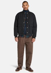 Zwarte gewatteerde bomberjack met twee voorzakken, gelaagd over een geruit shirt, gecombineerd met bruine broek en bruine veterschoenen.