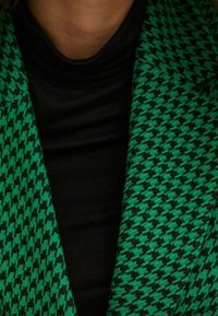 Blazer à motif pied-de-poule vert et noir fabriqué en tissu texturé, porté par-dessus un col roulé noir uni.