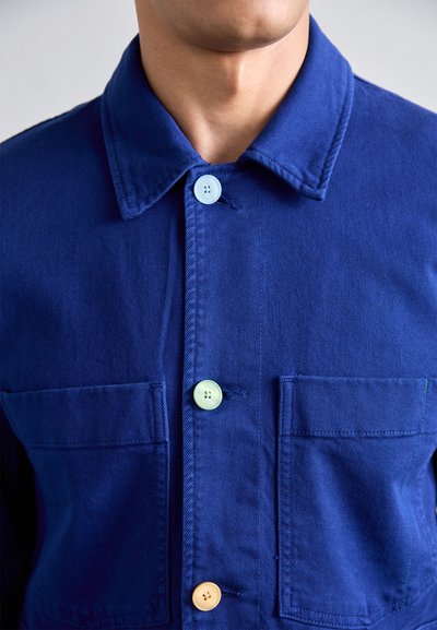 Blue denim jacket featuring a collared neck, two front pockets, and multicolored buttons in green, blue, and orange.