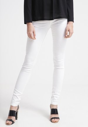 Osoba nosząca obcisłe białe spodnie i czarne sandały na wysokim obcasie, stojąca z lekko rozstawionymi nogami na jednolitym białym tle.