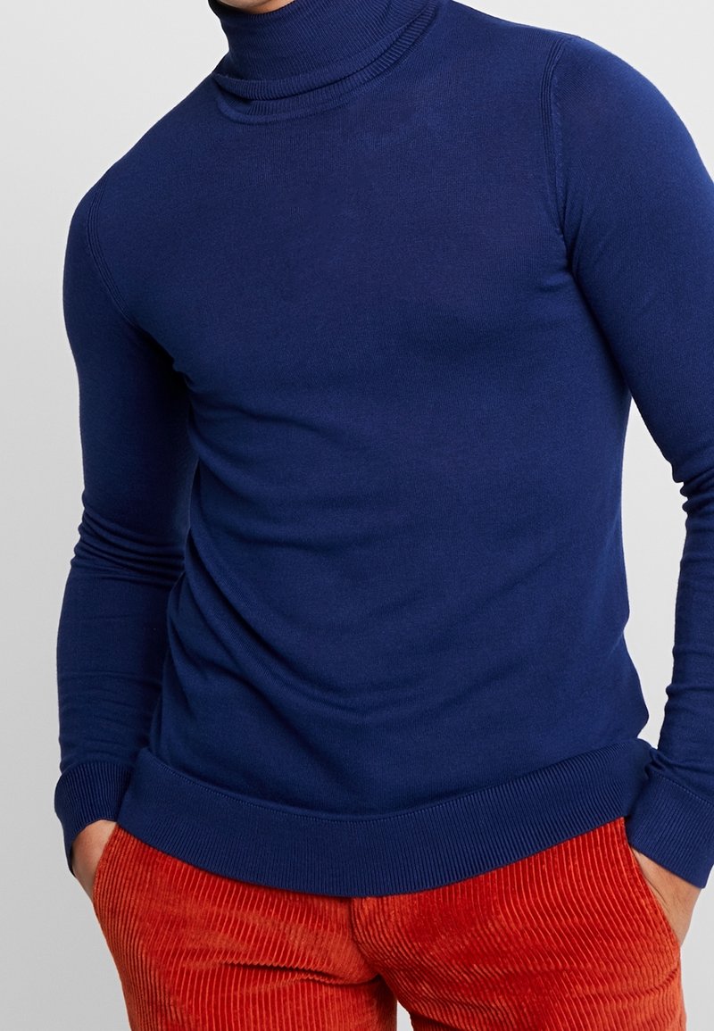 Mörkblå polotröja i ett slätt stickat tyg med långa ärmar och en figurnära passform, kombinerad med strukturerade orange manchesterbyxor.