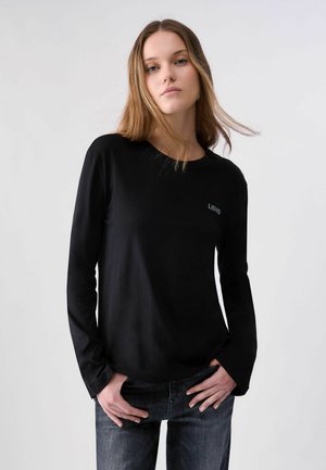 Donna con capelli lunghi indossa una semplice maglia nera a maniche lunghe e jeans scuri, in piedi con le mani in tasca contro uno sfondo chiaro.