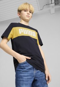 Donkerblauwe katoenen T-shirt met een gele horizontale streep waarop "PUMA" in witte tekst staat. Draag het met een blauwe denim jeans, hand in zak.