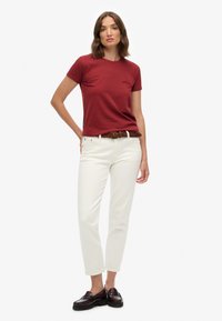 Bordeaux katoenen t-shirt, slim fit, korte mouwen; gecombineerd met witte rechte broek en een bruin gevlochten riem. Zwarte instappers maken de look compleet.