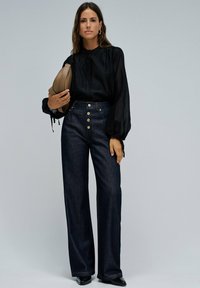 Donna che indossa una blusa nera a maniche lunghe e jeans blu scuro a vita alta, mentre tiene una borsa beige, in piedi davanti a uno sfondo semplice.