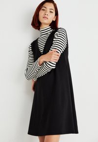 Vestido negro de corte A hecho de una tela suave, con un escote en V profundo, llevado sobre un cuello de tortuga a rayas en blanco y negro. Textura suave.