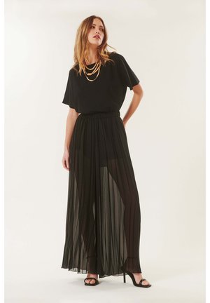 Femme debout portant un haut large noir, une jupe plissée noire transparente avec un short visible en dessous, des colliers dorés superposés et des sandales noires à talons hauts.