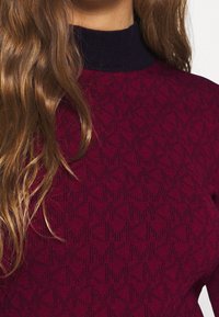 Pull bordeaux avec un motif texturé et un col côtelé bleu marine foncé. Tissu en maille, présentant des motifs géométriques subtils.