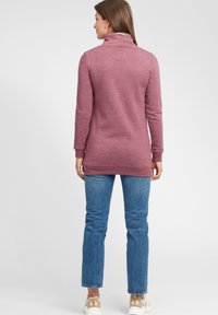 Burgundy tröja med hög krage, texturerad tyg och ett diskret logotypmärke på baksidan. Matchad med blå jeans och avslappnade sneakers.