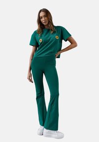 Zielona bawełniana koszulka i dzwonowate spodnie z haftem słonecznika na klatce piersiowej. Gładka tekstura, luźny krój, zestawione z białymi trampkami.