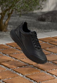 Zapatilla negra de cuero con detalles perforados, un upper texturizado y suela acolchada, posicionada sobre una superficie de azulejos con patrón.