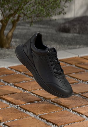Black leather sneaker with perforated details, a textured upper, and cushioned sole, positioned above a patterned tiled surface.