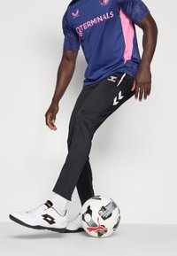 Un pantalon foncé ajusté, des chaussures de football blanches avec des accents noirs, et un maillot bleu et rose. Un ballon de football noir et blanc repose à côté du pied.