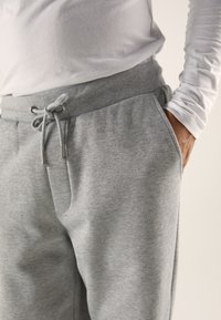 Gray sweatpants feature a drawstring waist, side pockets, and soft fabric with a smooth texture. Worn with a fitted white long-sleeve shirt.