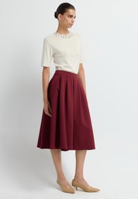 Haut crème avec un col orné, associé à une jupe midi bordeaux à plis. Porté avec des escarpins pointus de couleur chair.