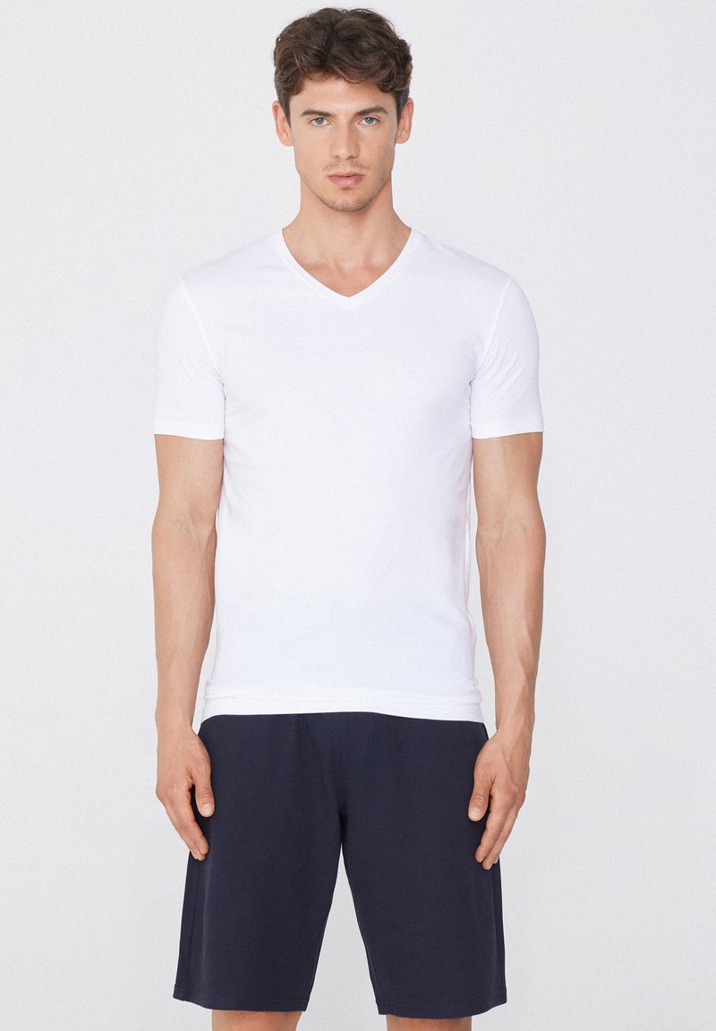 Joven de pie, vistiendo una camiseta blanca lisa de manga corta con cuello en V y pantalones cortos oscuros hasta la rodilla sobre un fondo blanco liso.