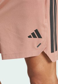 Shorts de sport rose clair en tissu texturé, avec des rayures latérales noires et un logo Adidas noir sur l'ourlet.