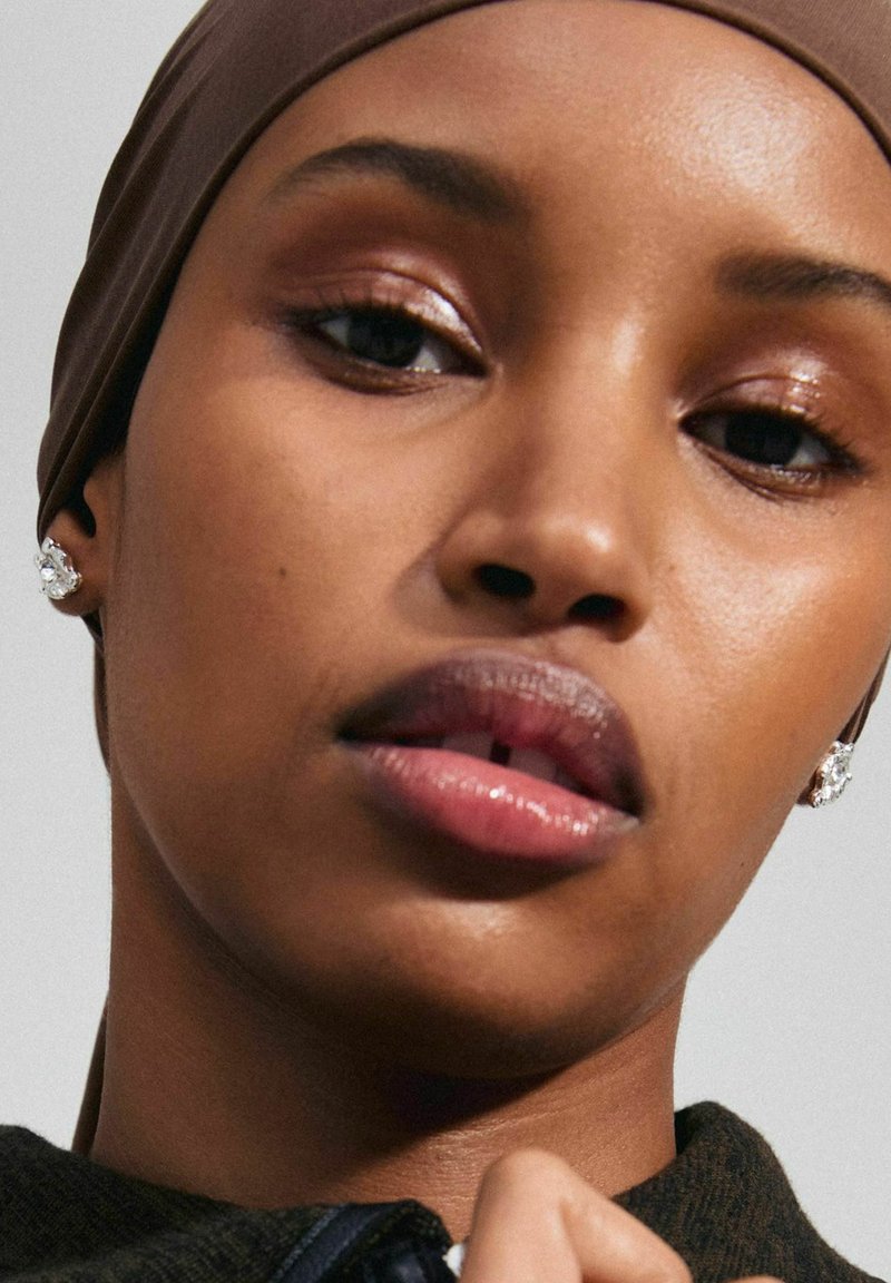Gros plan d'une personne portant un foulard brun, avec des lèvres brillantes, un maquillage subtil des yeux et des boucles d'oreilles en cristaux scintillants sur une peau lisse.