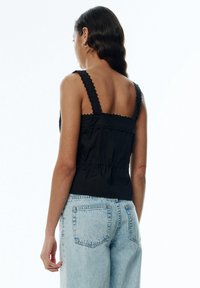 Débardeur noir avec bretelles festonnées, dos froncé et silhouette ajustée, associé à un jean denim bleu clair. Texture de tissu lisse.