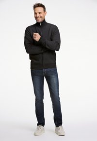 Sort, struktureret zip-jakke med høj krave; parret med mørkeblå jeans og hvide sneakers. Modellen står med armene korslagte og smiler.