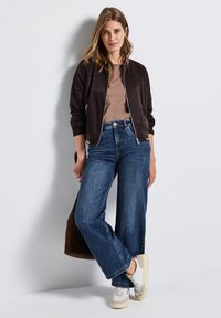 Veste aviateur marron sur un top beige, associée à un jean bleu à jambes larges et des baskets blanches. Un sac marron est tenu dans une main.