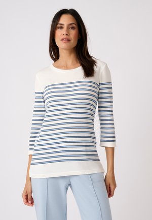 Femme aux cheveux foncés portant un haut blanc à manches longues avec des rayures horizontales bleues et un pantalon bleu clair, debout devant un fond uni.