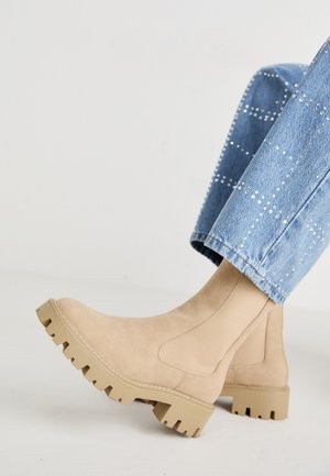 Beige semsket ankelstøvel med en kraftig gummisåle og elastiske paneler på siden, parret med lyseblå denim med naglede detaljer.
