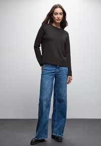 Dunkelbrauner Strickpullover mit einem gerippten Saum, kombiniert mit hellblauen weiten Jeans und schwarzen Schuhen, vor einem neutralen Hintergrund.
