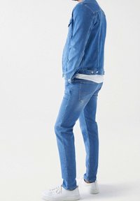 Muž ve modré džínové bundě a džínech s bílými teniskami, stojící bokem proti prostému bílému pozadí.