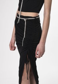 Falda negra con frunces y un cinturón trenzado plateado. Presenta aberturas laterales y cordones ajustables para un ajuste personalizable. Textura de tela suave.