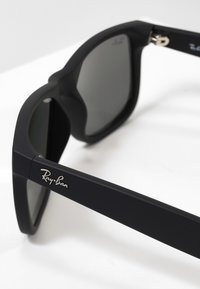 Svarta rektangulära solglasögon med gråa linser, matt finish, med silverfärgad Ray-Ban-logotyp på sidan. Robust båge och enkel design.