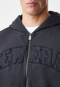 Felpa nera con zip caratterizzata da un logo "NEW ERA" cucito, materiale in cotone, polsini a costine e una cerniera frontale con un tirante metallico.