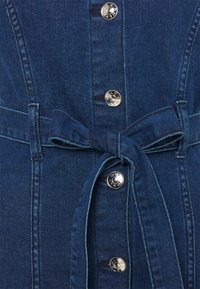 Mörkblå denimplagg med silverfärgade tryckknappar och ett knytband i samma tyg i midjan.