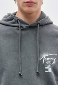 Νεαρός που φοράει γκρι φούτερ με κορδόνια, με λευκό κείμενο που αναφέρει πόλεις όπως το Λονδίνο, η Νέα Υόρκη, το Παρίσι, το Λος Άντζελες και το Τό κιο.