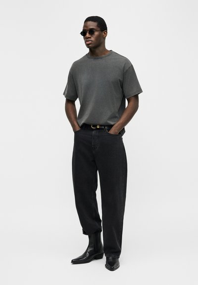 Άνδρας που φοράει σκούρα γυαλιά ηλίου, χαλαρή γκρι μπλούζα, μαύρη ζώνη, μαύρα τζιν σε χαλαρή εφαρμογή και μαύρα μποτάκια αστραγάλου, μπροστά σε απλό ανοιχτό φόντο.