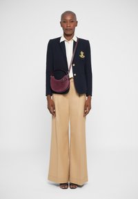 Άτομο που φοράει ναυτικό σακάκι με έμβλημα, λευκό πουκάμισο, φαρδιά μπεζ παντελόνια, μαύρες σαγιονάρες και κρατάει μια στρογγυλή σκούρα κόκκινη τσάντα στον ώμο.