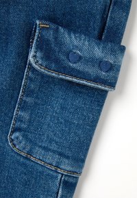 Tela de vaquero en azul medio, con un bolsillo con parche y acentos azules en forma de corazón, costuras visibles y una textura ligeramente desgastada.