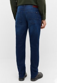 Donkerblauwe denim jeans met een rechte pasvorm, twee achterzakken, subtiele stikseldetails en een bruine leren patch op de taille.