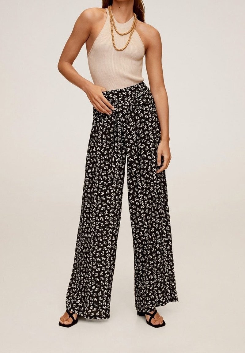 Femme portant un haut beige sans manches en côte, des colliers superposés en perles de bois, un pantalon noir à jambes larges avec imprimé floral blanc, et des sandales noires.