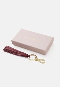 Porte-clés en cuir bourgogne avec un design de frange, quincaillerie en métal doré et un détail de logo. Accompagné d'une boîte rose clair avec des lettres dorées.