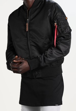 Man draagt een zwarte bomberjack met ritssluiting en een rood stoffen label aan het zakje op de arm, een zwart lang shirt en lichtgekleurde gescheurde spijkerbroek.