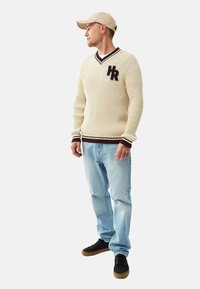 Krämfärgad ribbadvärmare med mörk vinröda accenter och broderad logotyp, tillsammans med ljusblå jeans och svarta sneakers. Beige keps.