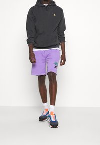 Sudadera con capucha negra con bolsillo canguro, pantalones cortos lavanda con un diseño impreso, y zapatillas multicolores con cordones blancos y detalles en blanco.