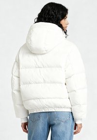 Doudoune blanche à capuche, avec des sections matelassées horizontales, des poignets élastiques et une coupe décontractée. Associée à un jean bleu clair.