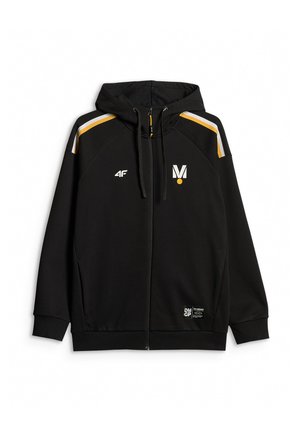 Młody mężczyzna w czarnej odzieży sportowej z logo, czapka z daszkiem, stoi na jednolitym szarym tle, zwrócony twarzą do przodu z neutralnym wyrazem.