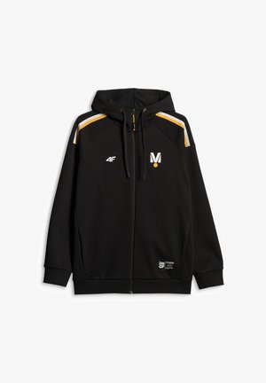 Młody mężczyzna w czarnej odzieży sportowej z logo, czapka z daszkiem, stoi na jednolitym szarym tle, zwrócony twarzą do przodu z neutralnym wyrazem.