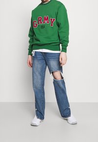 Felpa verde con logo "GRMY", colletto e polsini a coste; abbinata a jeans leggeri blu con strappi alle ginocchia e bottoni a pressione.