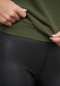 Olivengrønn skjorte som trekkes litt opp for å avsløre svarte leggings. Skjorten har en myk tekstur, mens leggingsene sitter tett og er glatte.