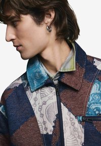 Kurtka z kołnierzem o wzorze patchworkowym w odcieniach niebieskiego, brązu i z teksturami w wzory. Zapięcie na zamek błyskawiczny oraz wyraźne detale kołnierza.